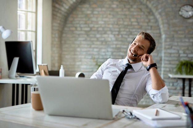 Un hombre de negocios feliz usando auriculares mientras se comunica con alguien y usa una computadora en la oficina