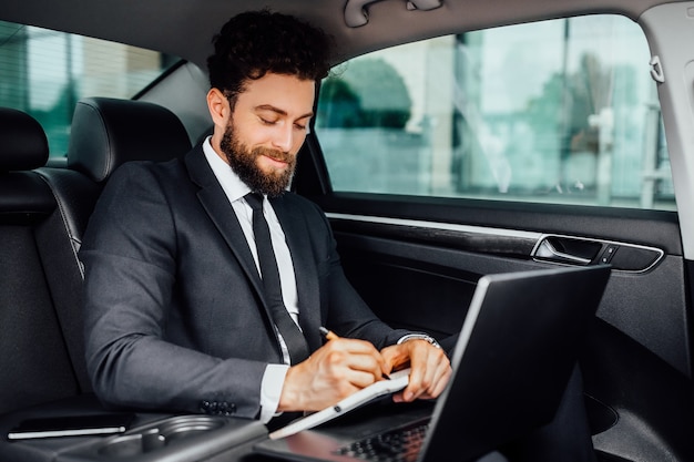 Hombre de negocios guapo, barbudo y sonriente que trabaja en el asiento trasero del automóvil y toma notas en el cuaderno desde su computadora portátil