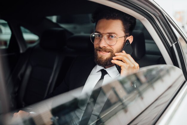 Hombre de negocios guapo, barbudo y sonriente en traje negro llamando por teléfono en el asiento trasero del coche