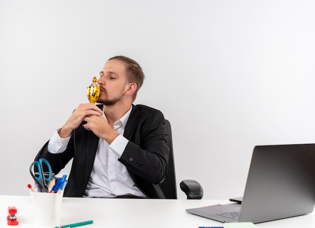 Hombre de negocios guapo complacido en traje besando su trofeo sentado en la mesa en offise sobre fondo blanco.