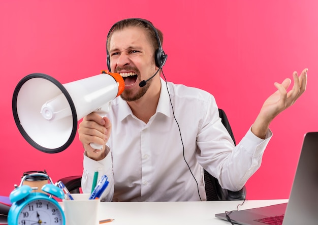 Hombre de negocios guapo enojado con camisa blanca y auriculares con un micrófono gritando al megáfono con expresión agresiva sentado en la mesa en offise sobre fondo rosa