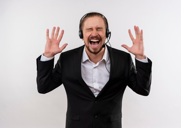 Hombre de negocios guapo frustrado en traje y auriculares con un micrófono gritando y gritando con expresión agresiva de pie sobre fondo blanco.