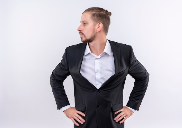 Hombre de negocios guapo molesto vestido con traje mirando a un lado de pie sobre fondo blanco.