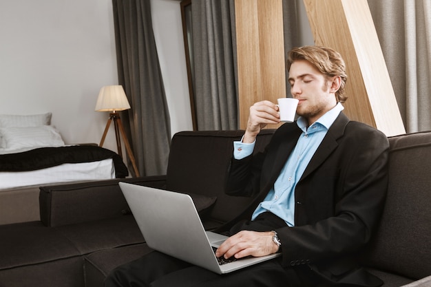 Hombre de negocios guapo relajado con peinado sylish y barba sentado en la habitación del hotel, tomando café, trabajando en un nuevo proyecto de inicio. Lugar de trabajo cómodo