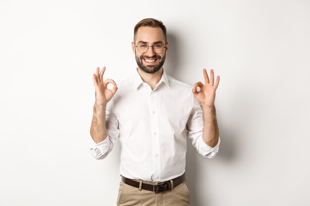 Hombre de negocios guapo satisfecho que muestra el signo de ok, calidad de garantía, de pie satisfecho contra el blanco