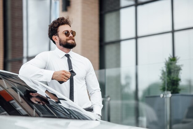 Hombre de negocios guapo, sonriente, feliz y barbudo está usando su teléfono móvil y está parado cerca de su automóvil al aire libre en las calles