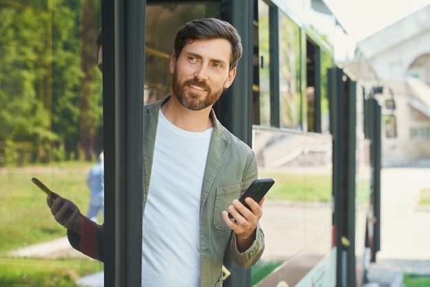 Hombre de negocios guapo sonriente con teléfono celular bajando del transporte público vista frontal de barbudo ocupado