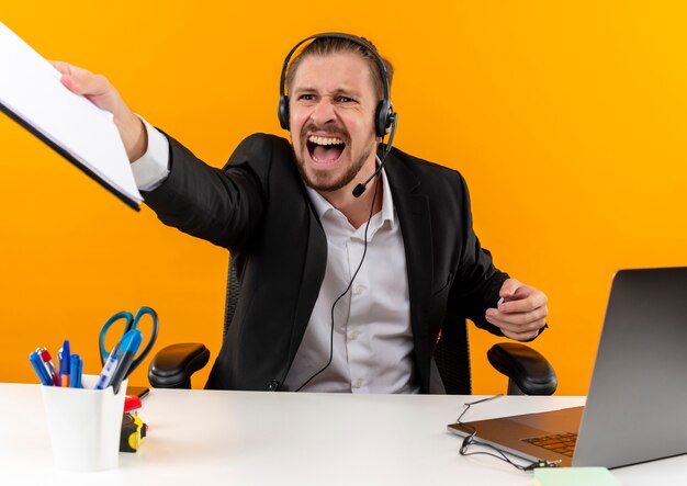Hombre de negocios guapo en traje y auriculares con un micrófono sosteniendo portapapeles mirando a un lado gritando con expresión agresiva sentado en la mesa en offise sobre fondo naranja