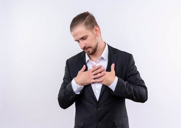 Hombre de negocios guapo vestido con traje cogidos de la mano en el pecho sintiendo emociones positivas y agradecidas con los ojos cerrados de pie sobre fondo blanco