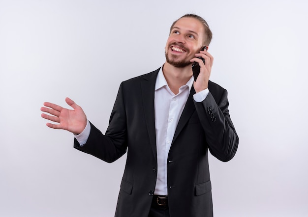 Hombre de negocios guapo vestido con traje hablando por teléfono móvil sonriendo de pie sobre fondo blanco.