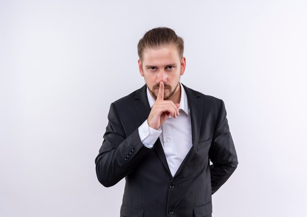 Hombre de negocios guapo vestido con traje haciendo gesto de silencio con el dedo en los labios sobre fondo blanco.