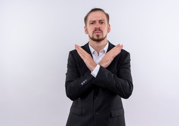 Hombre de negocios guapo vestido con traje haciendo señal de stop cruzando los brazos mirando a la cámara con rostro serio de pie sobre fondo blanco.