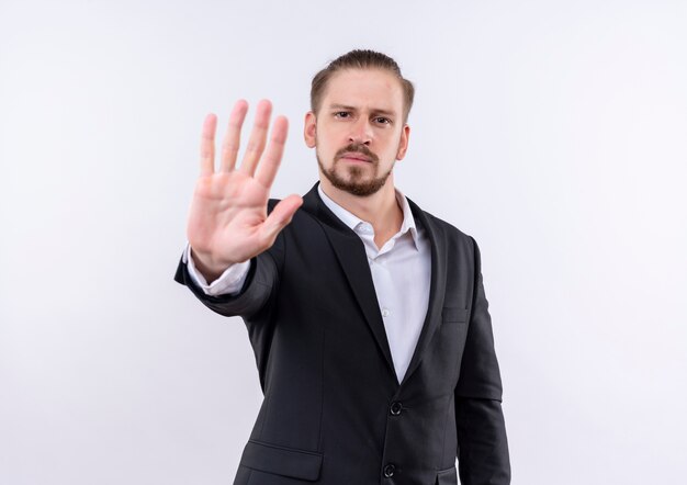 Hombre de negocios guapo vestido con traje haciendo señal de stop con la mano abierta mirando a la cámara con rostro serio de pie sobre fondo blanco.