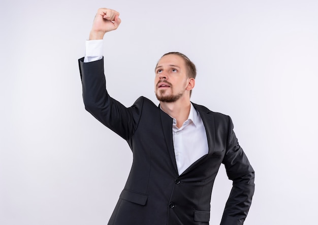 Hombre de negocios guapo vestido con traje mirando hacia arriba mostrando el puño con cara seria de pie sobre fondo blanco.