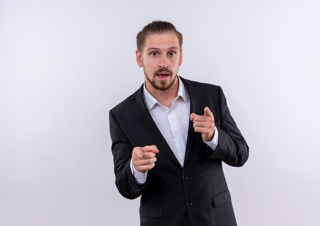 Hombre de negocios guapo vestido con traje mirando a la cámara apuntando con el dedo índice a la cámara de pie sobre fondo blanco.