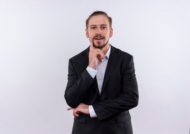 Hombre de negocios guapo vestido con traje mirando a la cámara con expresión pensativa pensando en posición positiva sobre fondo blanco.