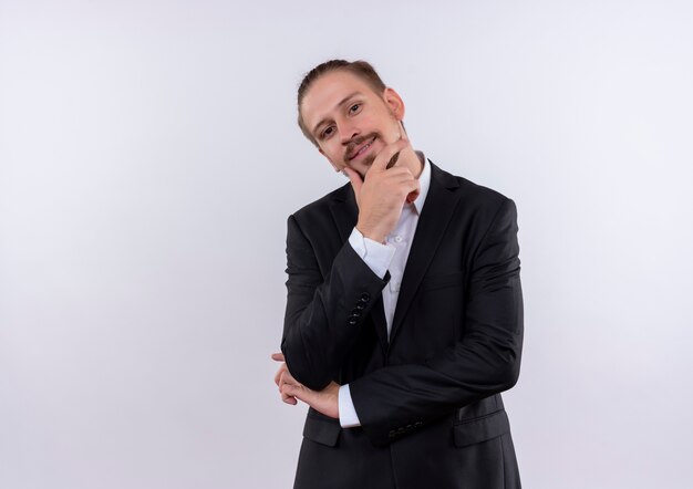 Hombre de negocios guapo vestido con traje mirando a la cámara con expresión pensativa pensando en posición positiva sobre fondo blanco.