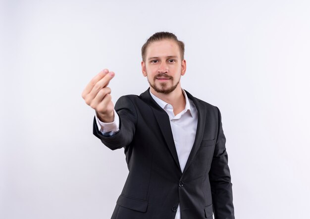 Hombre de negocios guapo vestido con traje mirando a la cámara frotando los dedos haciendo gesto de dinero de pie sobre fondo blanco.