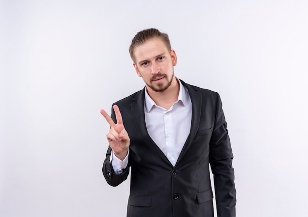 Hombre de negocios guapo vestido con traje mirando a la cámara sonriendo confiado mostrando signo de victoria de pie sobre fondo blanco.