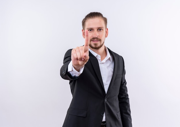 Hombre de negocios guapo vestido con traje mirando a la cámara sonriendo mostrando el dedo índice de pie sobre fondo blanco.