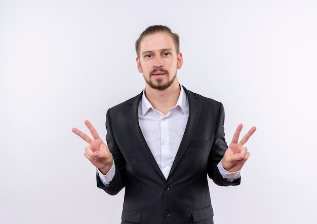 Hombre de negocios guapo vestido con traje mirando a la cámara sonriendo mostrando el signo de la victoria con ambas manos de pie sobre fondo blanco.