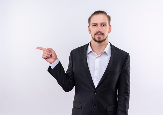 Hombre de negocios guapo vestido con traje mirando a la cámara con una sonrisa en la cara apuntando con el dedo hacia el lado de pie sobre fondo blanco.