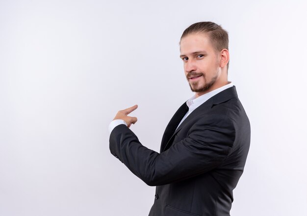 Hombre de negocios guapo vestido con traje mirando a la cámara con sonrisa de confianza apuntando hacia atrás de pie sobre fondo blanco.