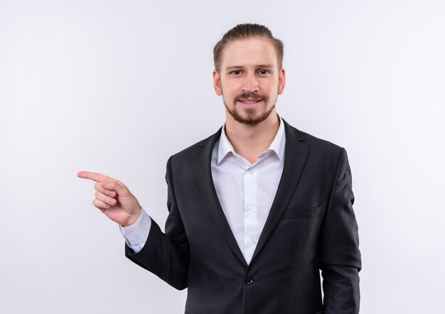 Hombre de negocios guapo vestido con traje mirando a la cámara con sonrisa de confianza apuntando con el dedo índice hacia el lado de pie sobre fondo blanco.