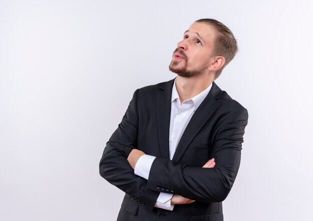 Hombre de negocios guapo vestido con traje mirando a un lado con expresión pensativa de pie sobre fondo blanco.