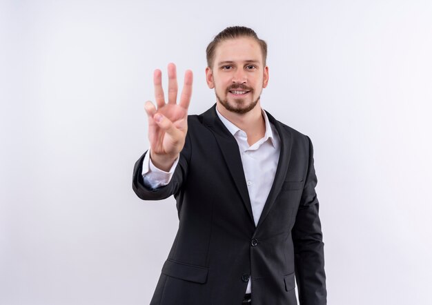 Hombre de negocios guapo vestido con traje mostrando y apuntando hacia arriba con los dedos número tres de pie sobre fondo blanco.