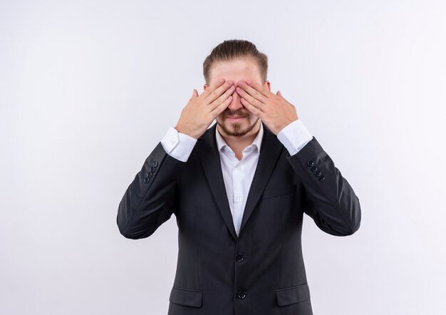 Hombre de negocios guapo vestido con traje que cubre los ojos con las manos de pie sobre fondo blanco.