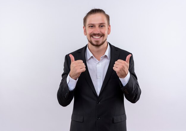 Hombre de negocios guapo vestido con traje sonriendo con cara feliz mostrando los pulgares para arriba de pie sobre fondo blanco.