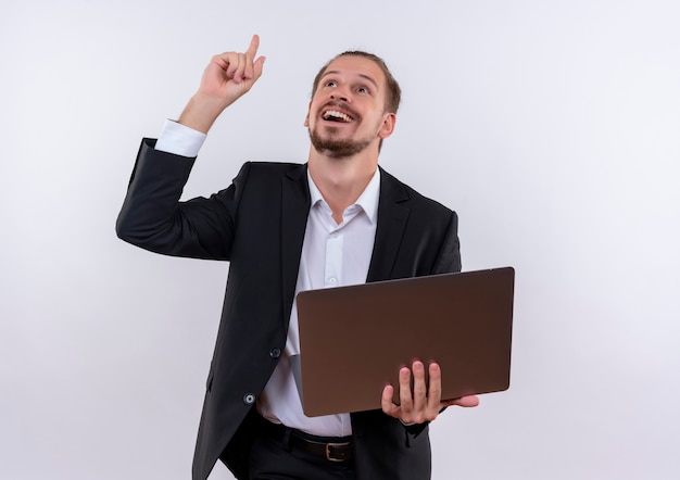 Hombre de negocios guapo vestido con traje sosteniendo la computadora portátil apuntando hacia arriba con el dedo sonriendo alegremente de pie sobre fondo blanco.