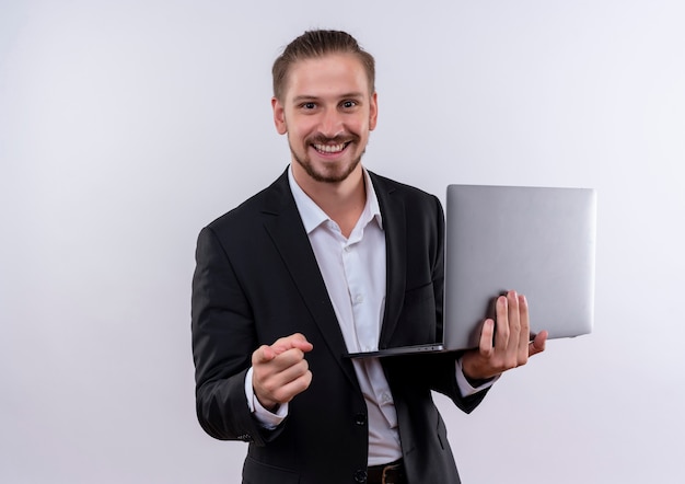 Hombre de negocios guapo vestido con traje sosteniendo la computadora portátil apuntando con el dedo a la cámara sonriendo alegremente de pie sobre fondo blanco.