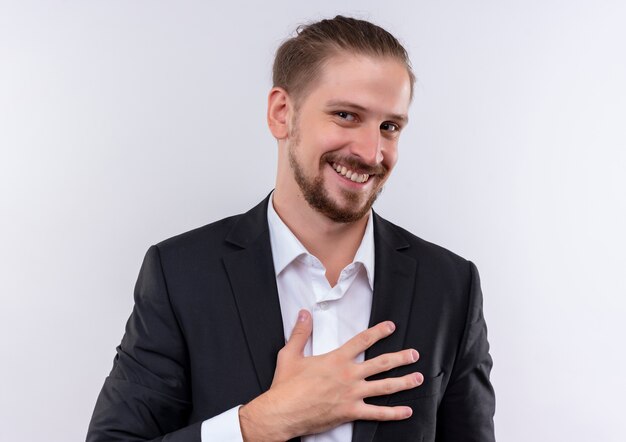 Hombre de negocios guapo vestido con traje sosteniendo la mano en el pecho mirando a la cámara con una sonrisa en la cara de pie sobre fondo blanco.