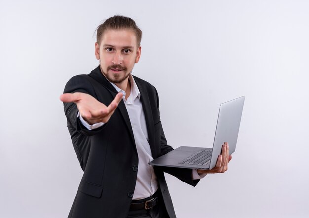 Hombre de negocios guapo vestido con traje sosteniendo el ordenador portátil con el brazo mirando a la cámara con una sonrisa en la cara de pie sobre fondo blanco.