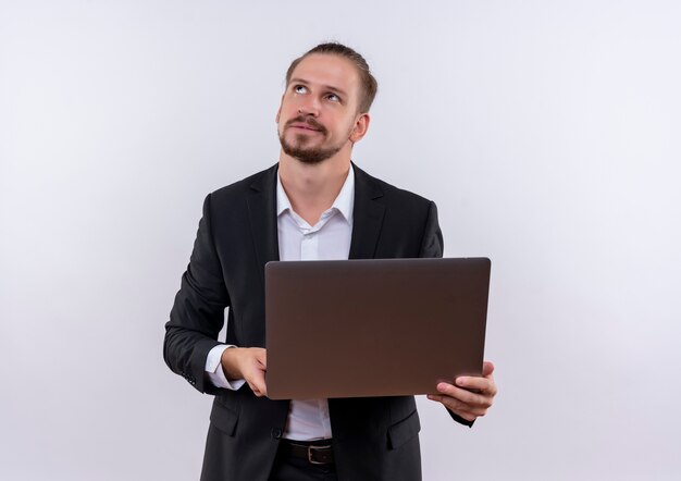 Hombre de negocios guapo vestido con traje sosteniendo ordenador portátil mirando hacia arriba con expresión pensativa de pie sobre fondo blanco.
