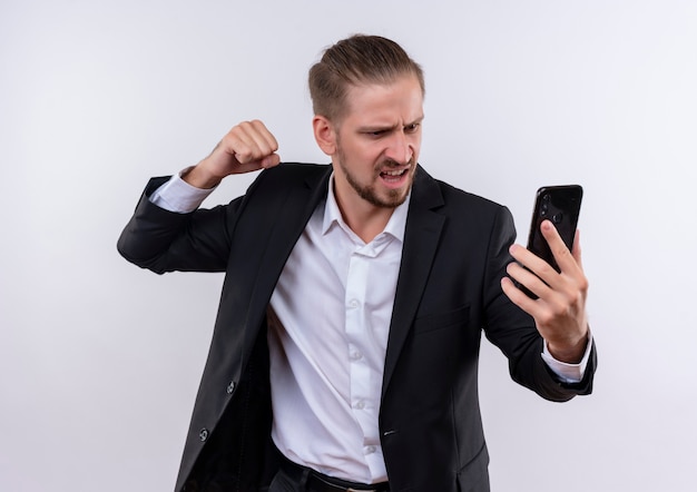 Hombre de negocios guapo vestido con traje sosteniendo smartphone mirando la pantalla con cara enojada con puño cerrado de pie sobre fondo blanco.
