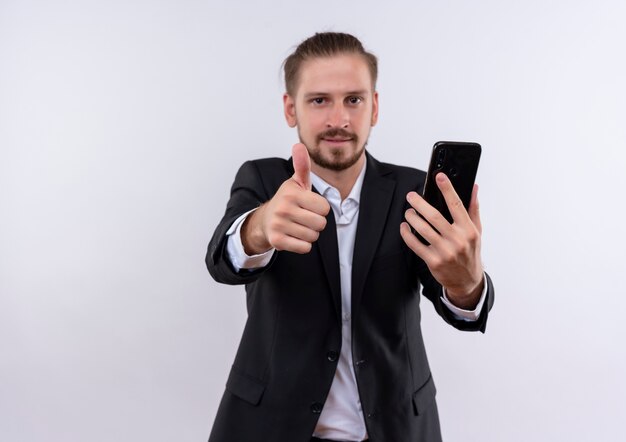 Hombre de negocios guapo vestido con traje sosteniendo smartphone sonriendo mostrando los pulgares para arriba de pie sobre fondo blanco.