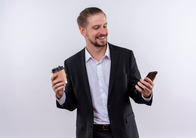 Hombre de negocios guapo vestido con traje sosteniendo smartphone y taza de café feliz y positivo de pie sobre fondo blanco.