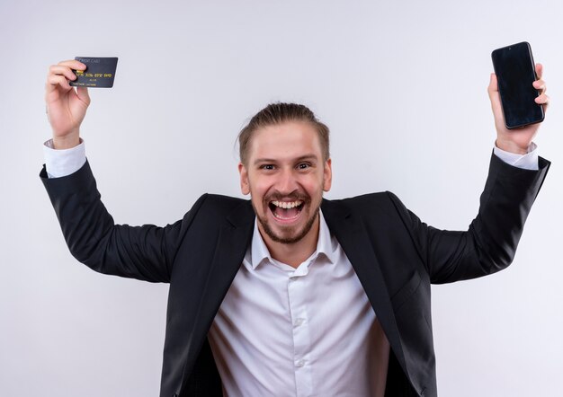 Foto gratuita hombre de negocios guapo vestido con traje sosteniendo teléfono inteligente y tarjeta de crédito en hads planteados feliz y emocionado de pie sobre fondo blanco.