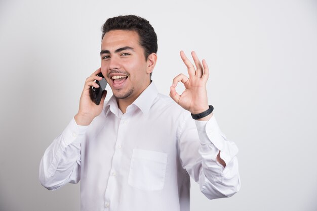 Hombre de negocios hablando con teléfono celular y mostrando signo ok sobre fondo blanco.