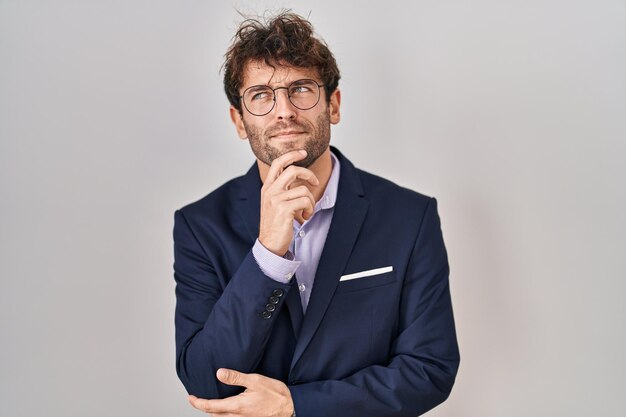 Hombre de negocios hispano con gafas con la mano en la barbilla pensando en la pregunta, expresión pensativa. sonriendo con cara pensativa. concepto de duda.