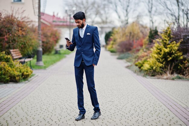 Hombre de negocios indio con barba elegante con bindi en la frente en traje azul posado al aire libre mirando el teléfono móvil