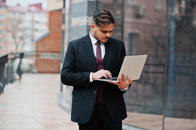 Hombre de negocios indio con estilo en ropa formal con portátil en las manos de pie contra las ventanas en el centro de negocios