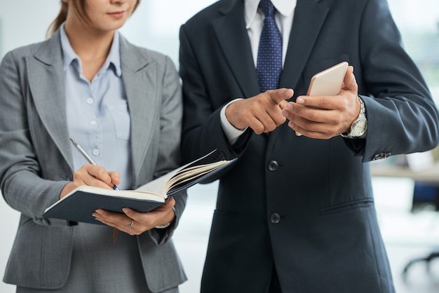 Hombre de negocios irreconocible en traje apuntando al teléfono inteligente en la mano, y una mujer tomando notas