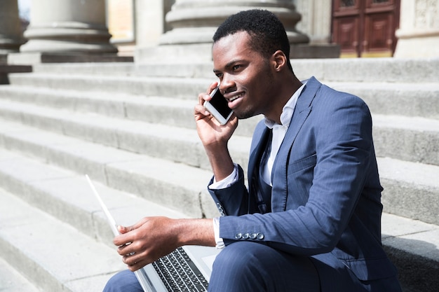 Hombre de negocios joven africano sonriente que habla en el teléfono móvil que se sienta en escalera con el ordenador portátil