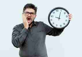 Foto gratuita hombre de negocios joven asustado con gafas sosteniendo el reloj de pared y poniendo la mano en la mejilla aislado en blanco