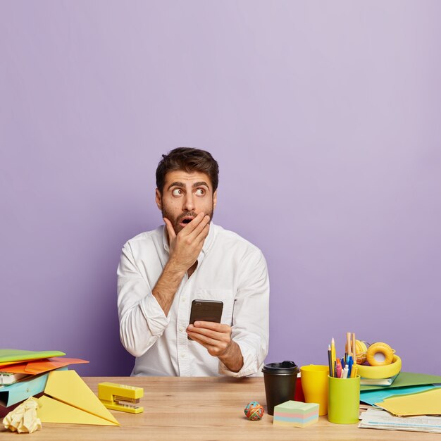 Hombre de negocios joven asustado mira arriba, sostiene el teléfono inteligente moderno, cubre la boca, sostiene el teléfono celular moderno en la mano, vestido con camisa blanca