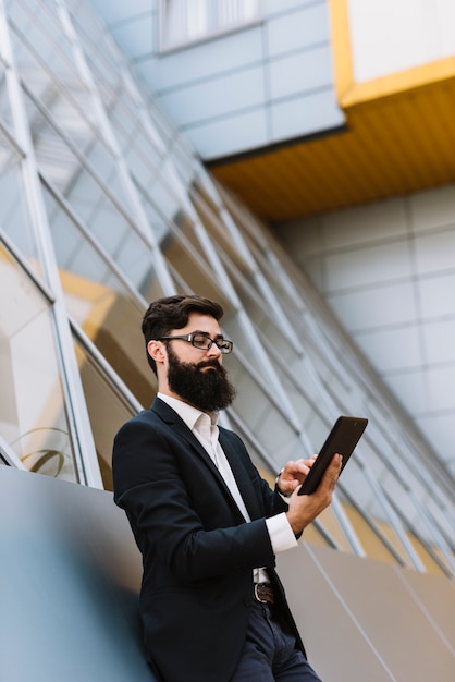 Foto gratuita hombre de negocios joven de la barba que se inclina en la pared que mira smartphone